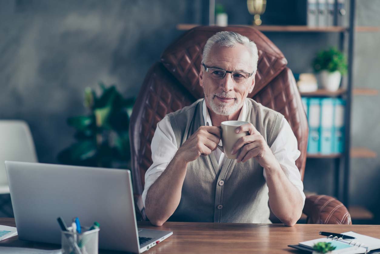 Retirement-age man sits at his work desk sipping coffee.